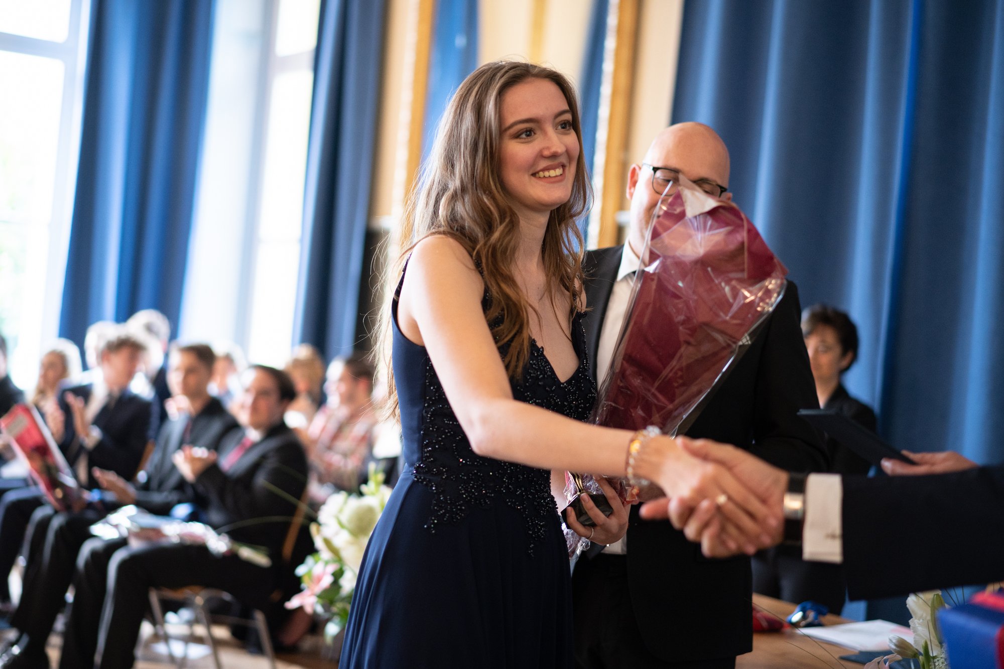 A female student shakes hand on a graduation day. Other students sit in the background. A female student shakes hand on a graduation day. Other students sit in the background.